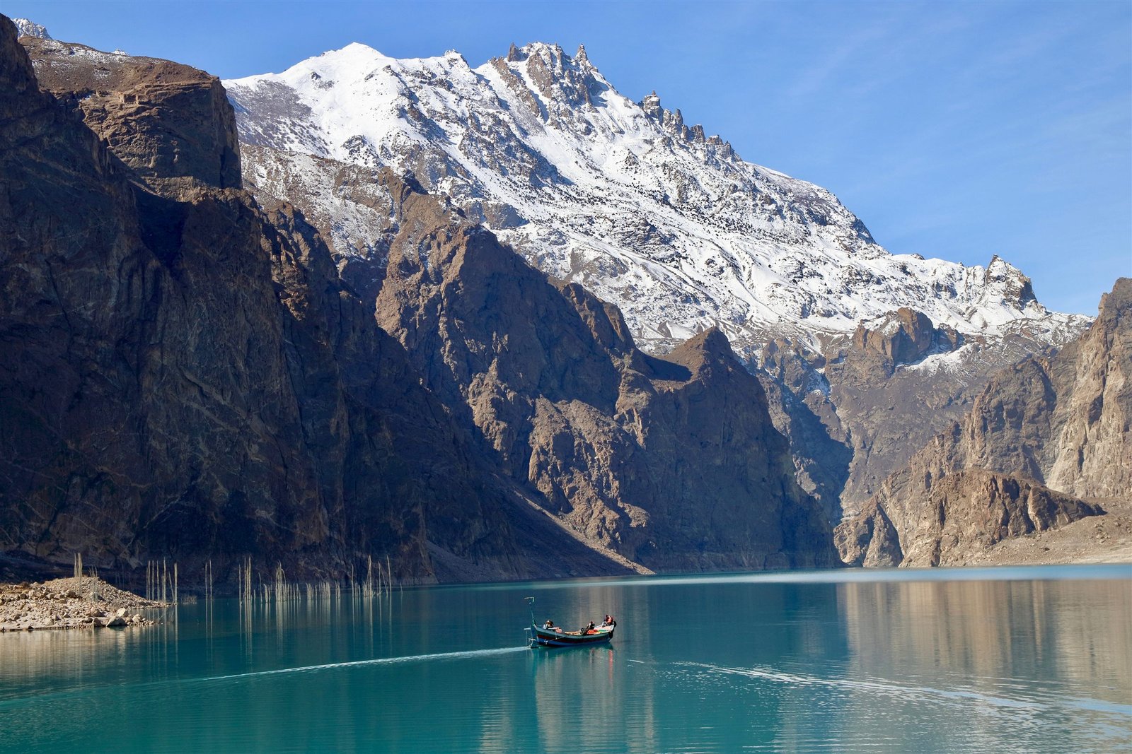 A wooden boat crossing Attabad Lake between steep rock walls and a snow peak