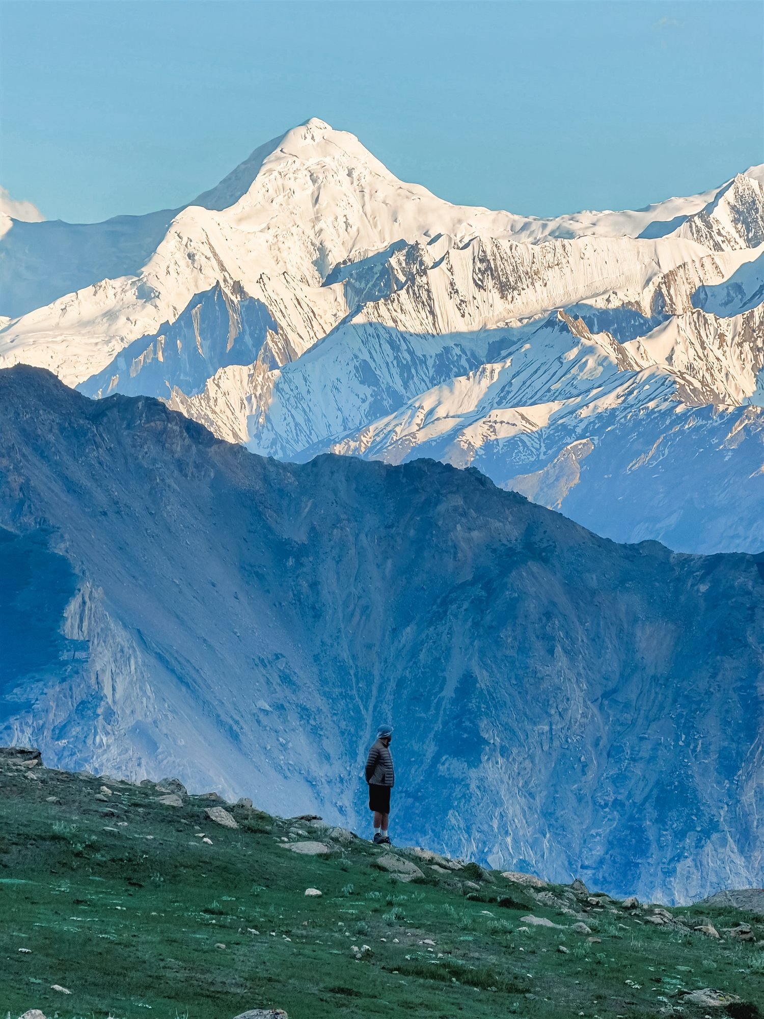 A hiker looking out over a deep valley toward a snow-capped peak at sunrise