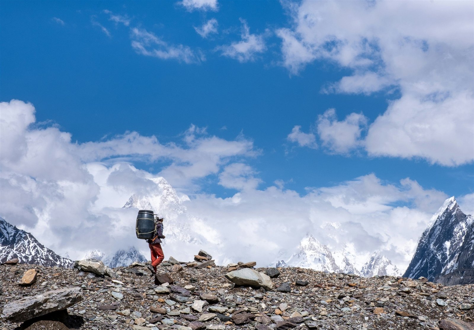 A porter carrying a load along the Baltoro moraine with K2 behind in cloud