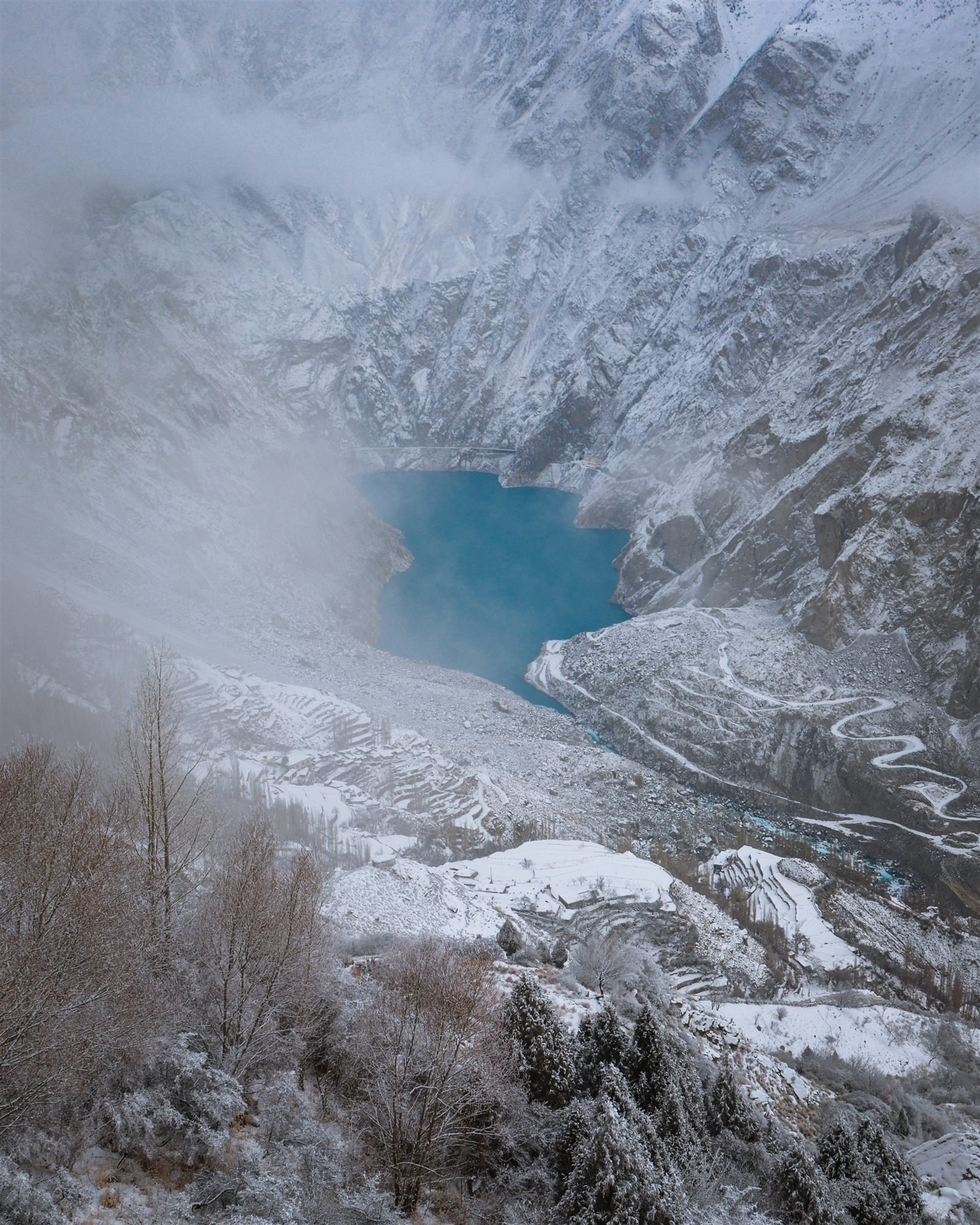 Snow and low cloud over Attabad Lake in winter, a switchback road cut into the slope